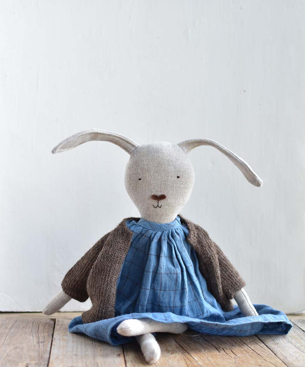 Ruthie le Rabbit, a fabric bunny doll in a brown sweater and blue dress, sits on a wooden floor against a plain white background.