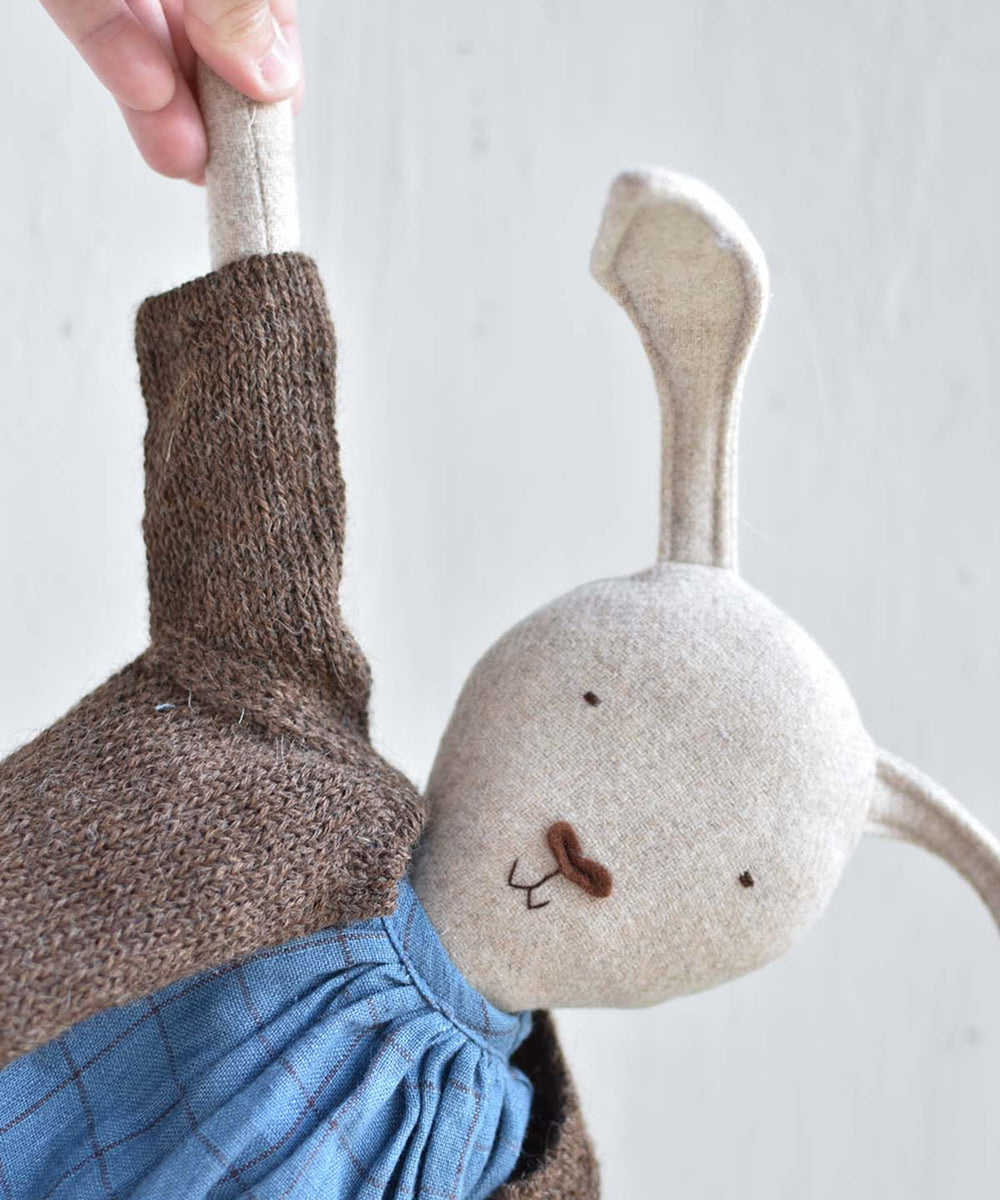 A hand holds Ruthie le Rabbit by the ear; she wears a blue dress and brown knitted sweater.