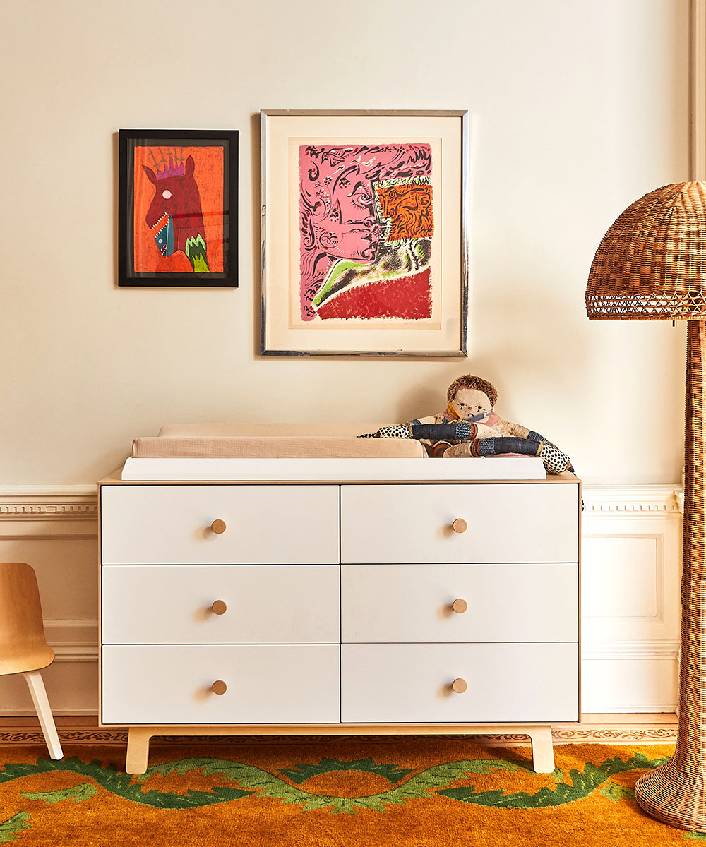 A white dresser with six drawers, a plush doll on top, artworks on the wall, a wicker lamp, and a wooden chair, set on a green and orange patterned rug.