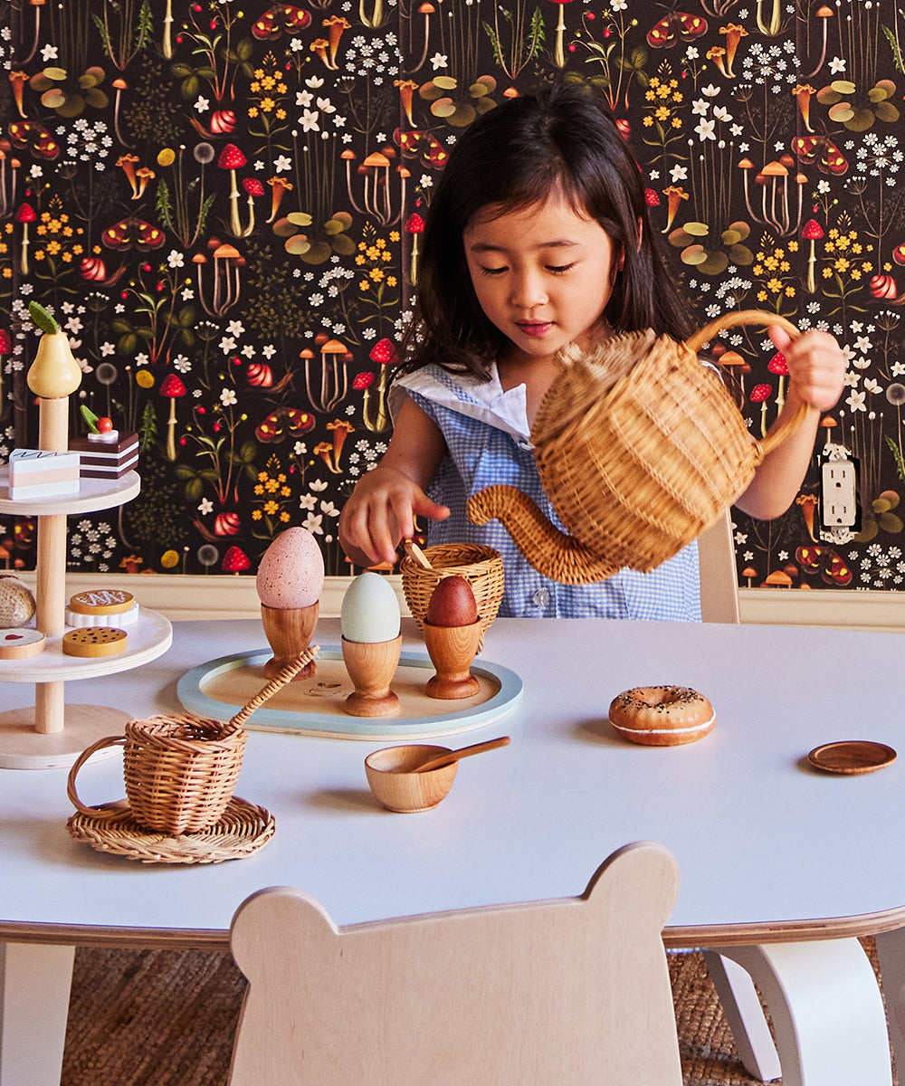 Young girl playing with the Le Wooden Dish Set at a small table, vibrant floral wallpaper in the background.