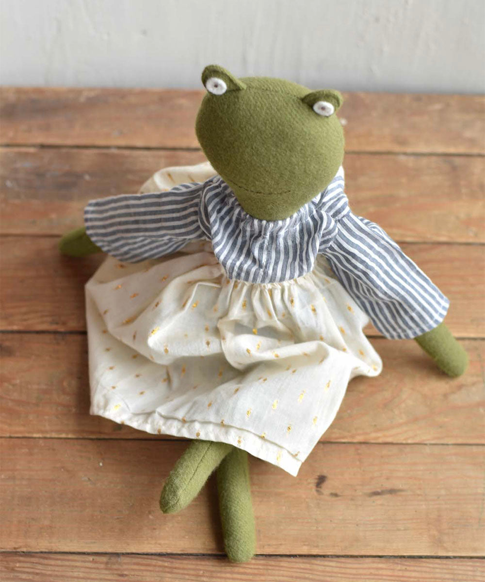 The Fern le Frog doll - Sparkle Skirt is a green stuffed frog wearing a striped shirt and white dress, seated on a wooden surface.