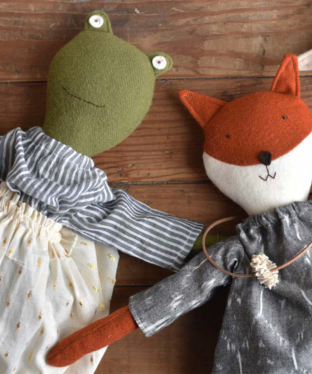 The Fern le Frog doll in a sparkle skirt lies on a wooden surface beside a fox doll, both handmade and dressed in cute outfits.