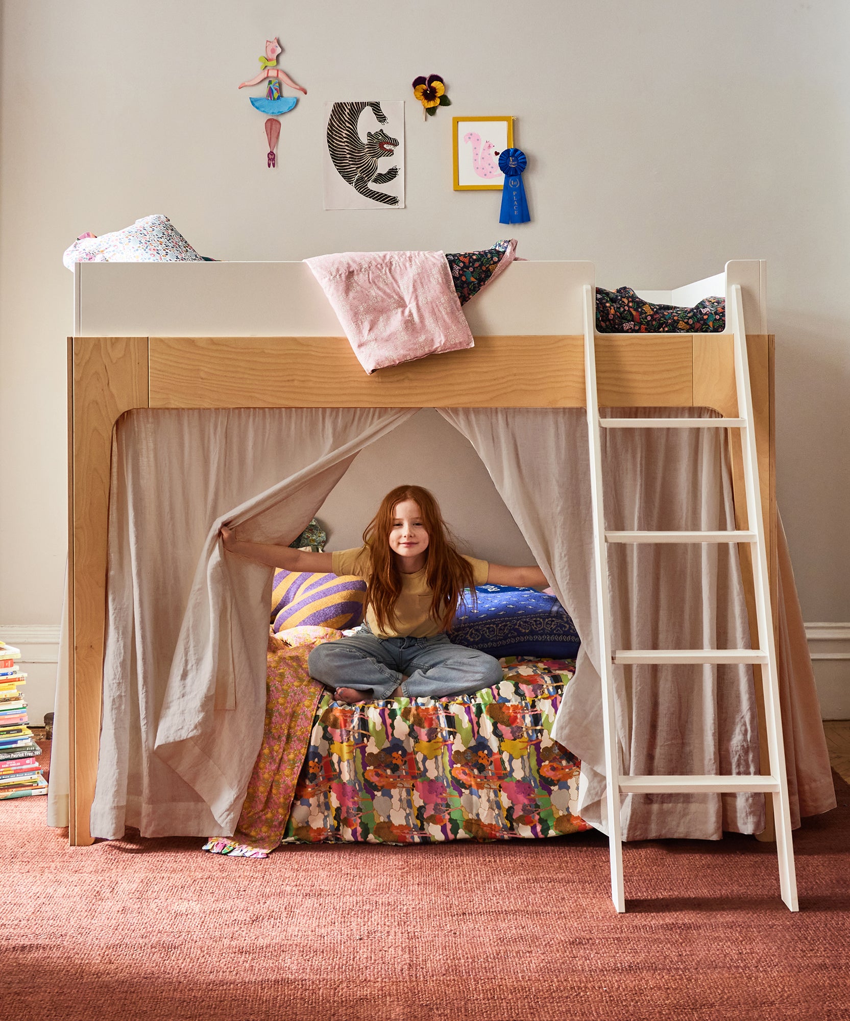 Young girl sits cross-legged on a colorful bed under a Perch Bunk Bed with Linen Curtains, surrounded by art and pillows.