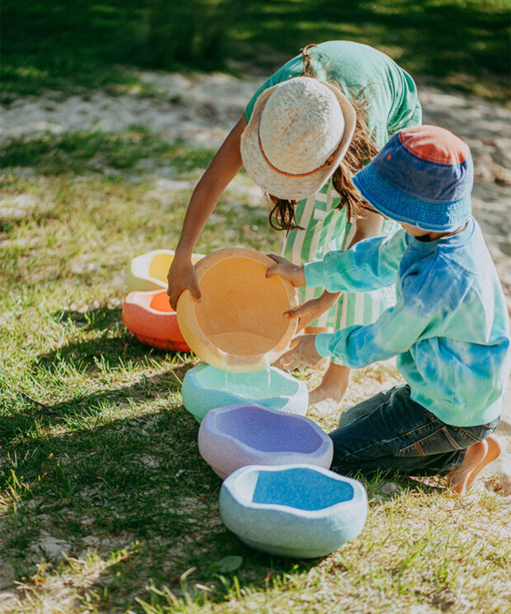 Two children in hats play outside, pouring water into Le Original Rainbow’s colorful, bowl-shaped containers on the grass.