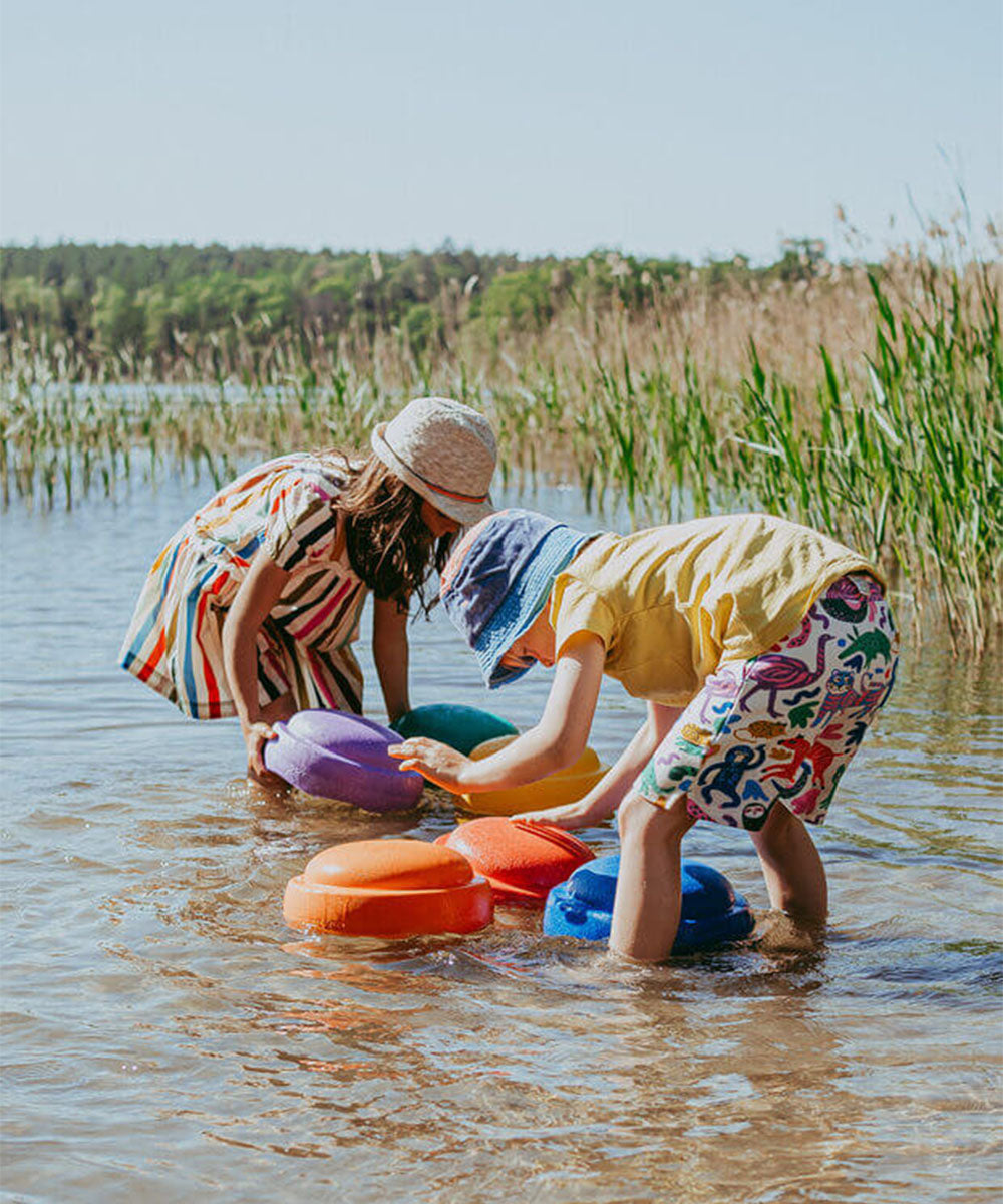 Two kids in sunhats play with Le Original Rainbow colorful discs in shallow water near tall grass on a sunny day.