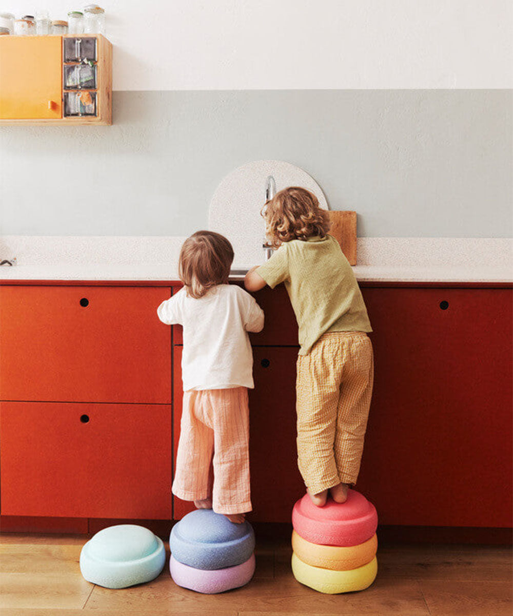 Two young children stand on Le Original Rainbow colorful cushions to reach a kitchen sink with red cabinets.