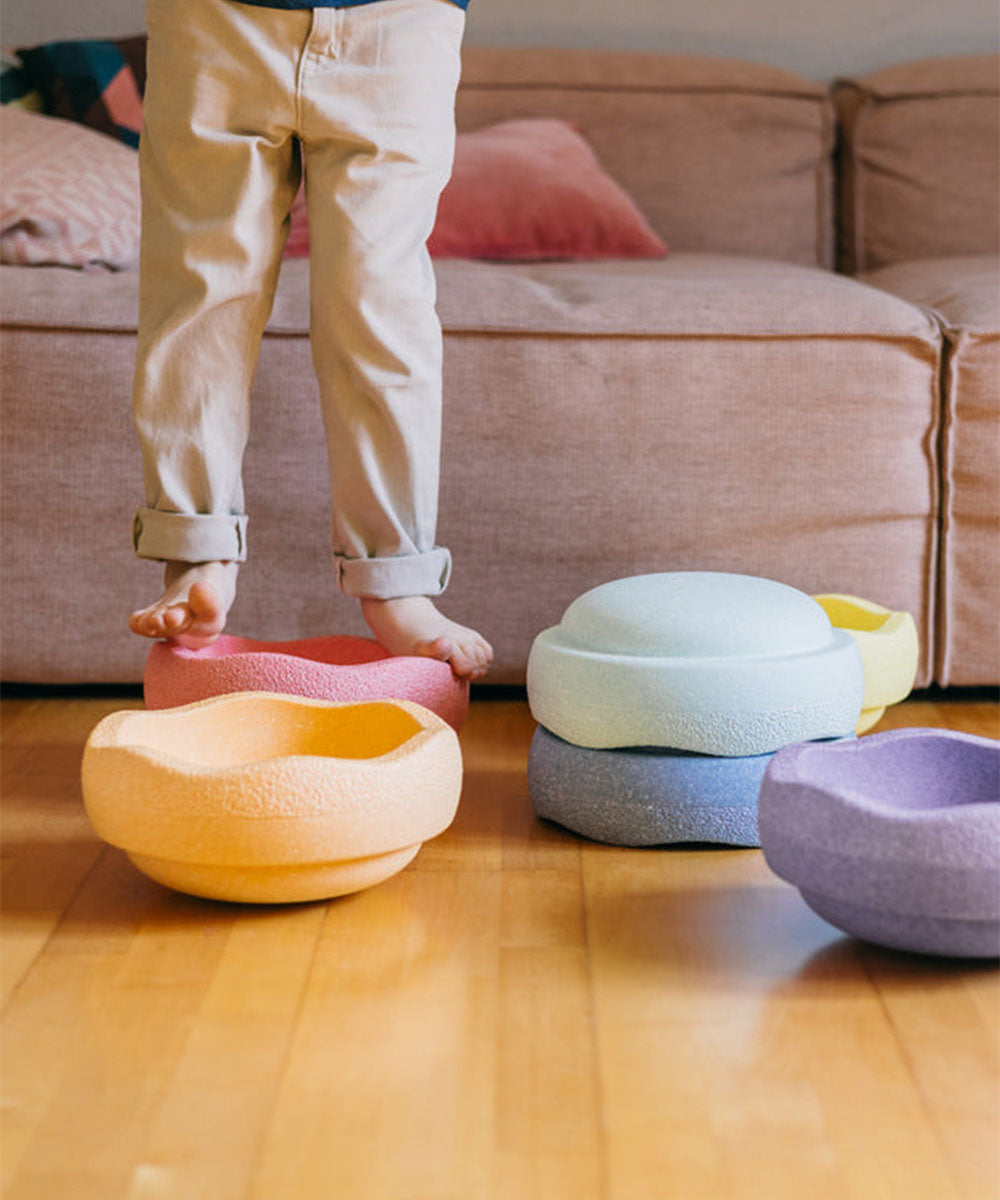 A child steps on Le Original Rainbow foam stepping stones in a living room with a beige sofa in the background.