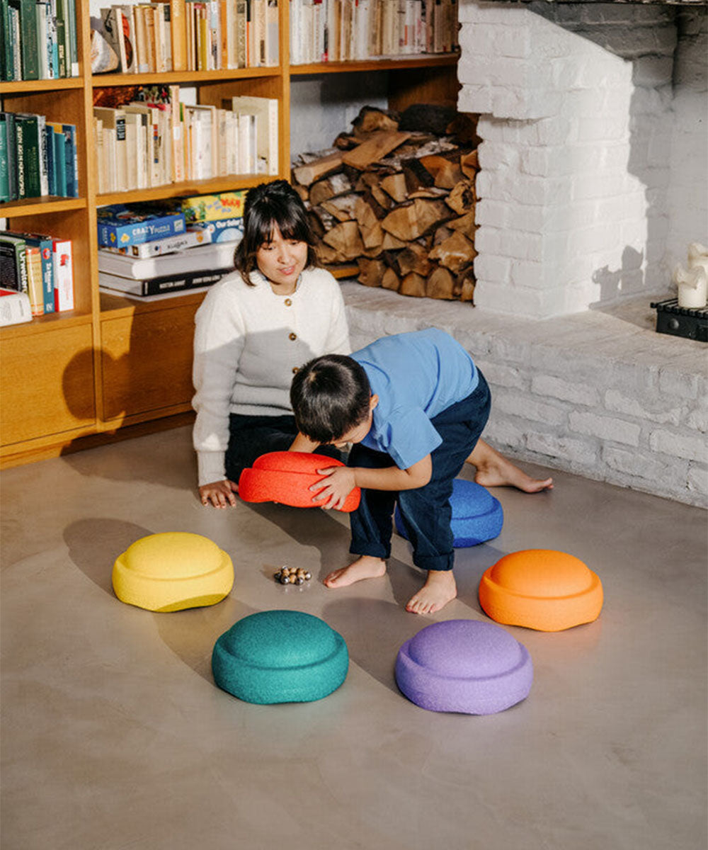 A woman and child play with the Le Original Rainbow colorful cushions on the floor in a cozy room with books and a fireplace.