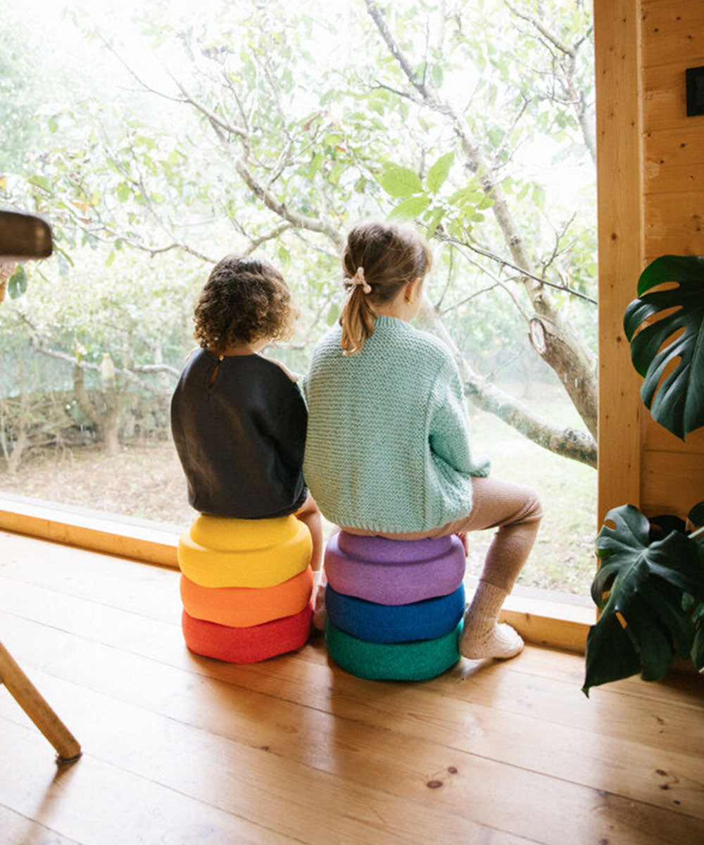 Two children sit on Le Original Rainbow stacked cushions, gazing out a large window at the trees and greenery outside.