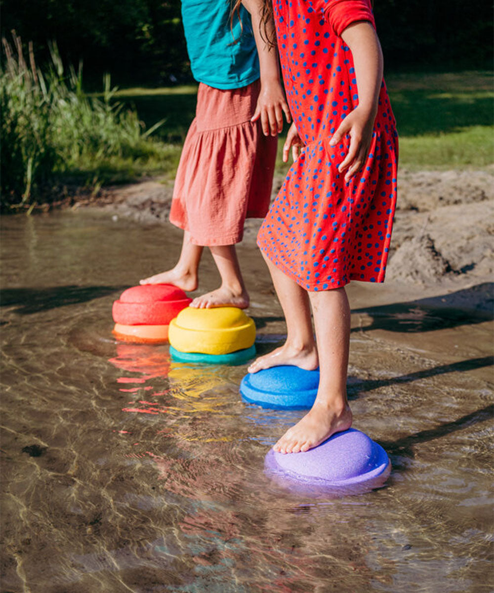 Two children balancing on Le Original Rainbow stepping stones in shallow water near the shore.