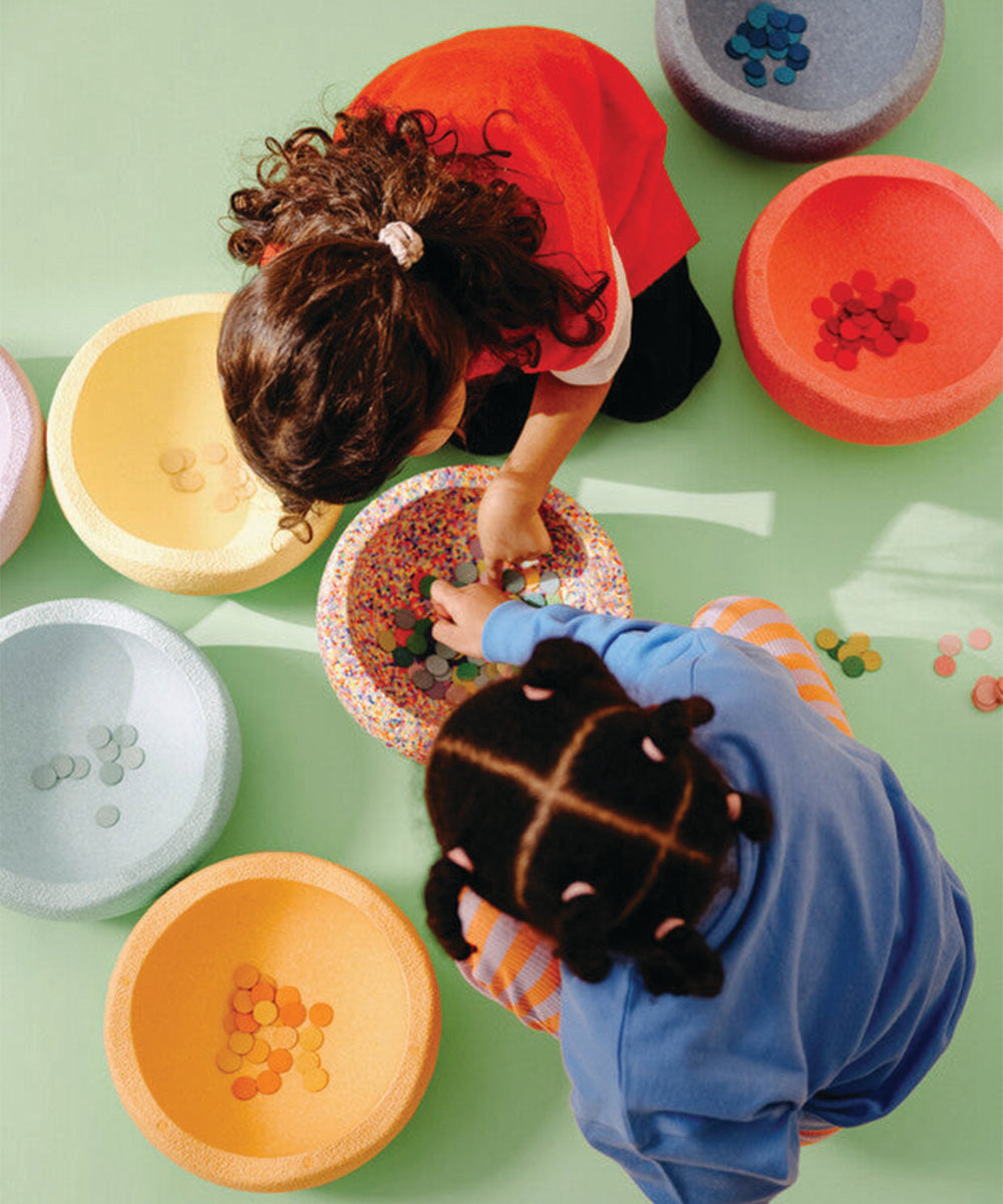 Two children sort the Complete Set's colorful discs into bowls on a light green surface, viewed from above.