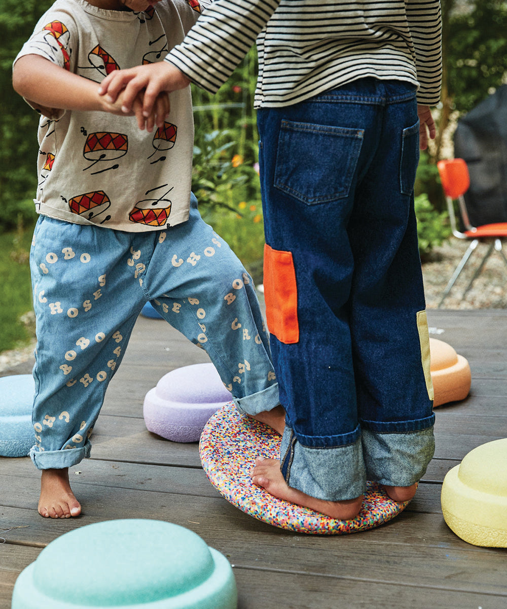 Two barefoot children balance on the Complete Set's colorful disc on a wooden deck, holding hands for support.
