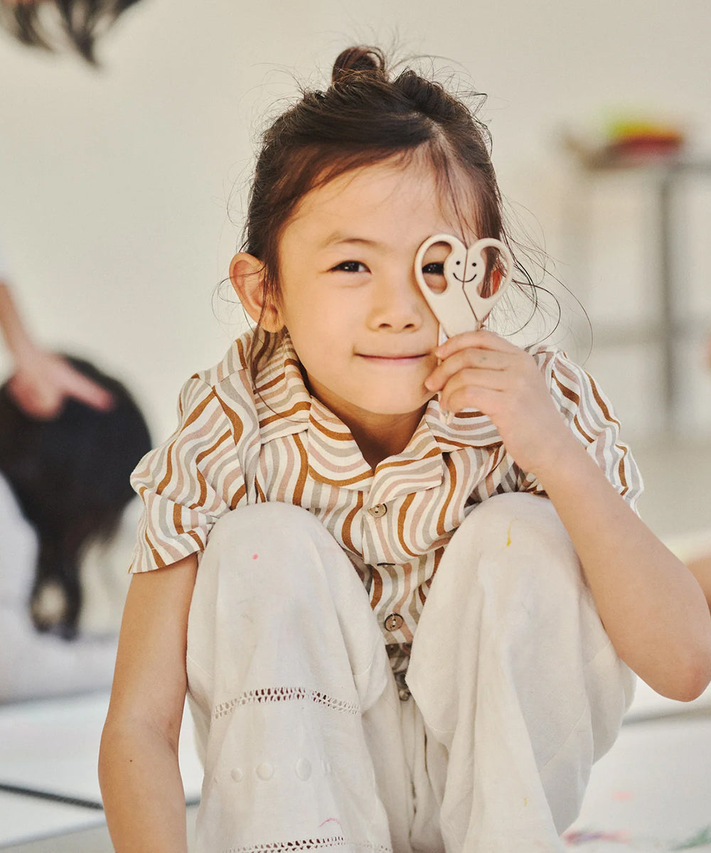 A young child smiles indoors while holding Le Gentle Scissors—playful, person-shaped scissors—up to their face.