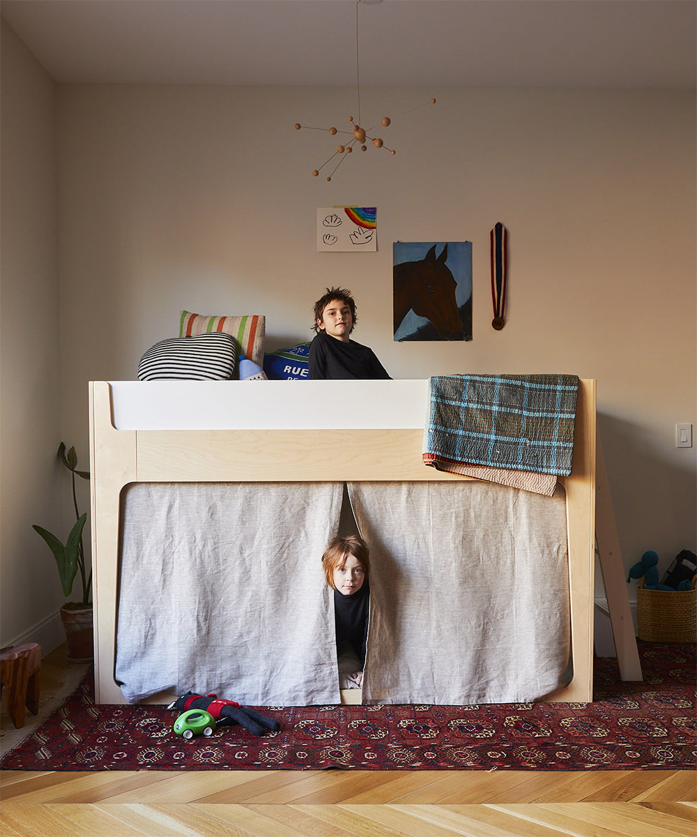 Nest loft bed, one boy under the loft peering through linen curtains attached to bed. Other boy sitting in the loft.