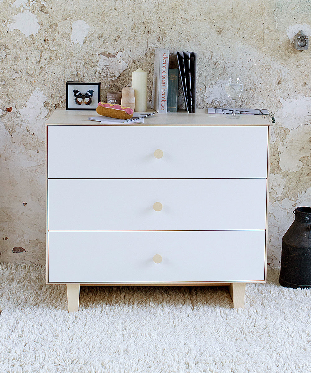 White and wood dresser with objects on top. Dresser has 3 deep drawers, light wood pulls and tapered wooden legs.